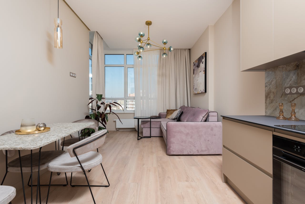 Interior of bright modern studio with open kitchen with cabinets near table and chairs next to couch with pillows near window with curtains
